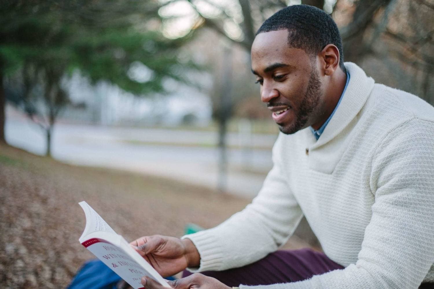 A man sitting outdoors, absorbed in reading something he is holding open in both hands.