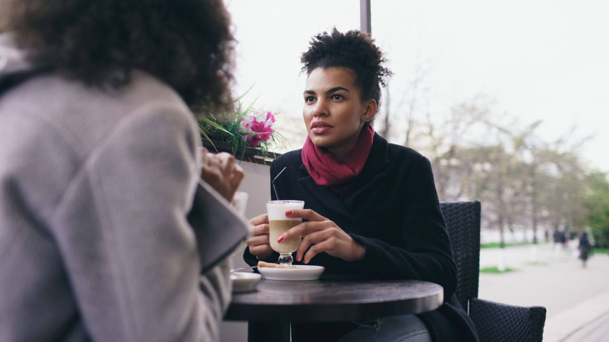 Two women talking across a small café table, one listening while the other speaks, a coffee between them.